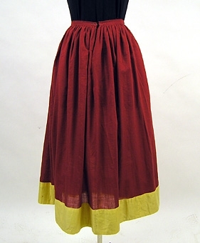 Kjol sydd av tunt, rött bomullstyg. Nederkanten dekorerad med ett gult satinvävt bomullstyg. Sprund mitt fram. Linningen knäppes med hyska och hake.

Enl liggare: 100570:1-4 "Västgötadräkt av bomull, bestående av vit blus, rött livstycke, röd kjol samt grönt förkläde."

"Brukats av Lizzie Lindgren som möjligen sytt den på Vara folkhögskola 1922"