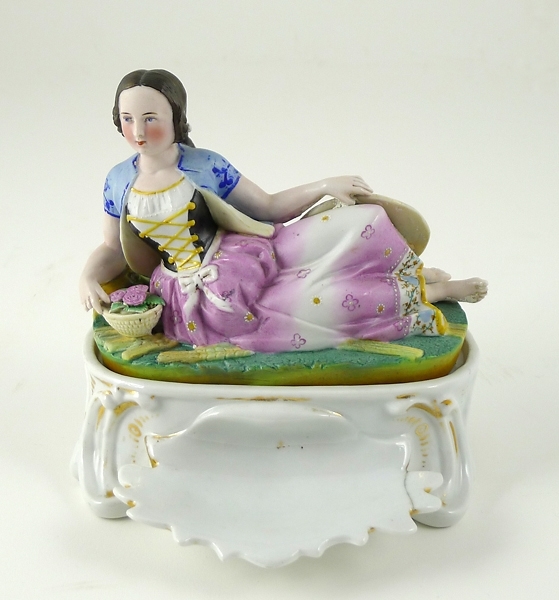 Enl. liggare: "Skrivställ av porslin, 1800-tals-rokoko, på locket en liggande kvinna."