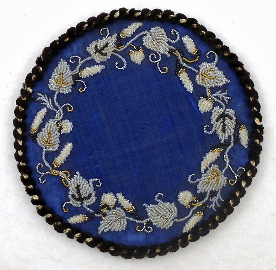 Lampmatta, rund, av blå sammet med ranka av pärlor i vitt, guld och svart samt snodd i svart och vitt silke runt kanten. Stadig papp inuti samt grå flanell på baksidan.