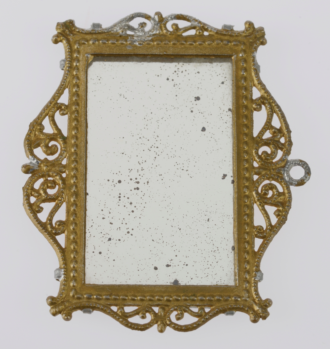 Et lite speil med ramme av gullforgylt metall. Metallet er ganske mykt. Rammen har dekor i form av snirklete mønster. Glasset er festet med fire små metallklemmer. Speilet har antageligvis vært i et dukkehus.
