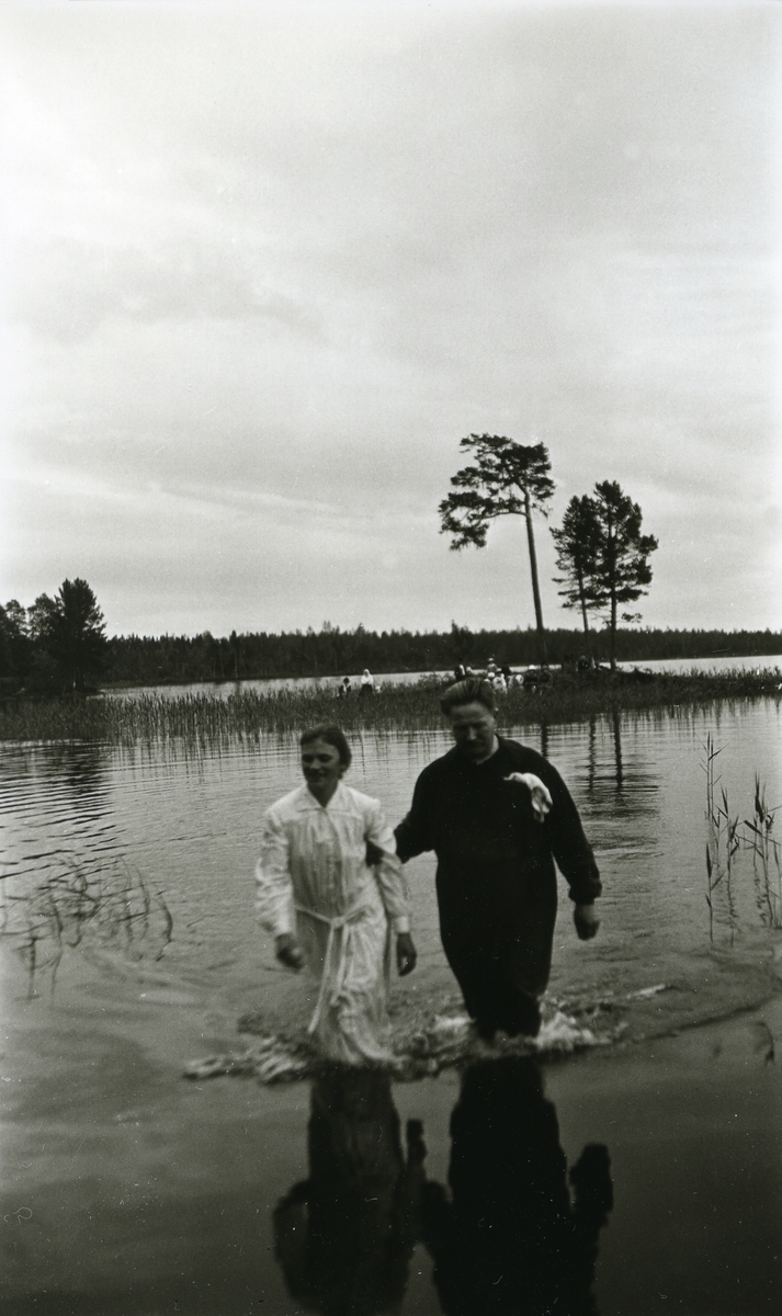 Presten og en av de døpte på vei opp fra vannet i Älvdalen finnmark, Sverige. Mennesker på holmen i bakgrunnen.