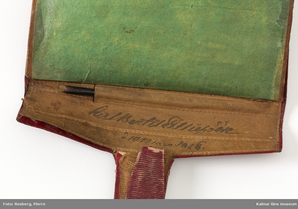 KLM 22667:1. Plånbok av läder och papper. Röd saffian med grönt pappersfoder, två fack. Namn skrivet med bläck inuti: "Carl Bertil Lilliehöök d. 10 mars 1828". Innehåller en tunn blyertspenna och ett fragment av en skrapnos-pinne, av elfenben. Ett av facken sönder på den ena kortsidan.