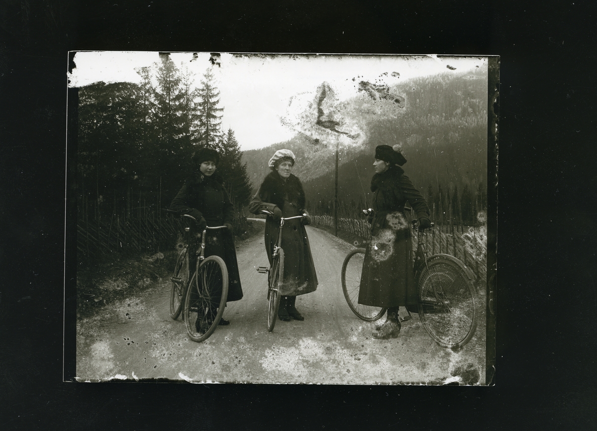 3 kvinner, i lange kåper står ved hver sin sykkel på veien ved Øylo i Vang.