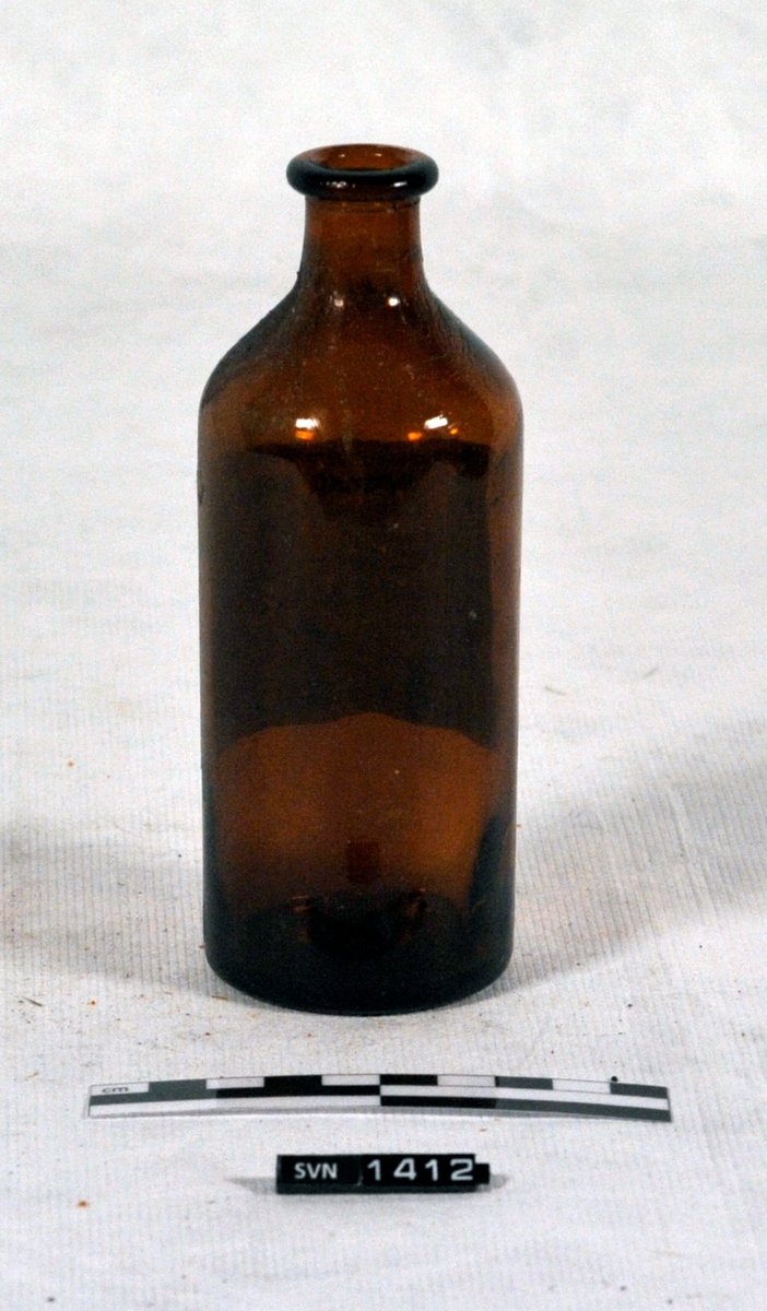 Brun flaske med hvit etikett med håndskrevet innholdsbeskrivelse. Etiketten har trykket logo og navn.
Flasken er tom og har ikke kork