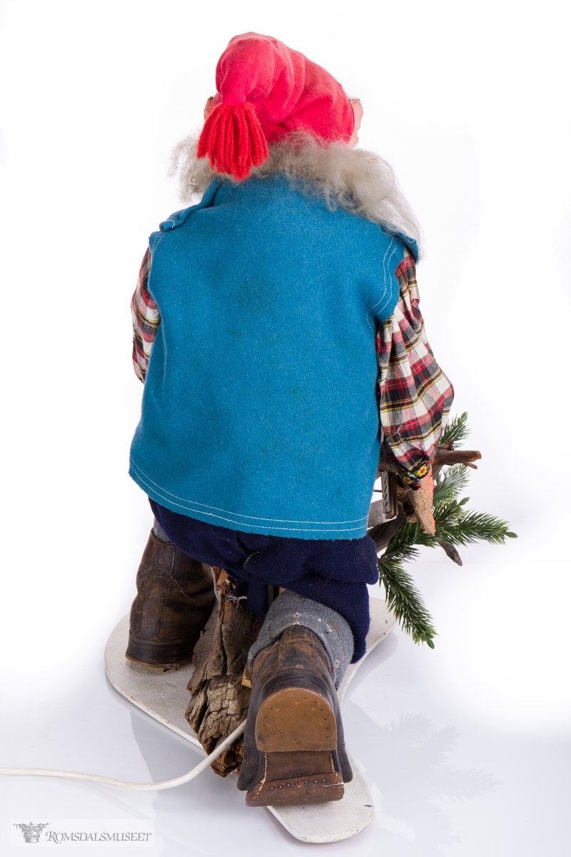 Mekanisk juledekorasjon bestående av en nisse som sitter på en fururot med en grindsag. Nissen er kledd i skjorte, vamdelsvest, knebukser, knestrømper og beksømsko. Tilkoblet ledning med av og på knapp.