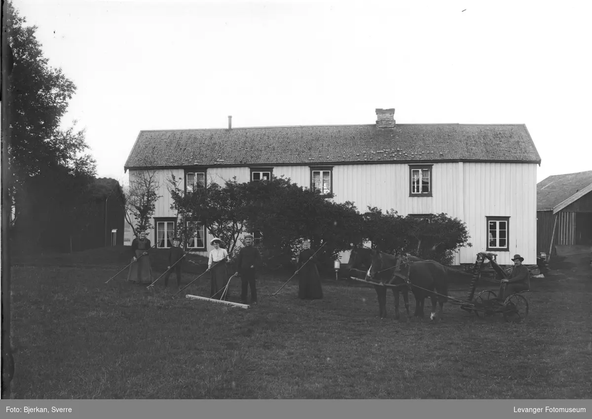 Familie fotografert med slåtteredskaper og hester i hagen.
