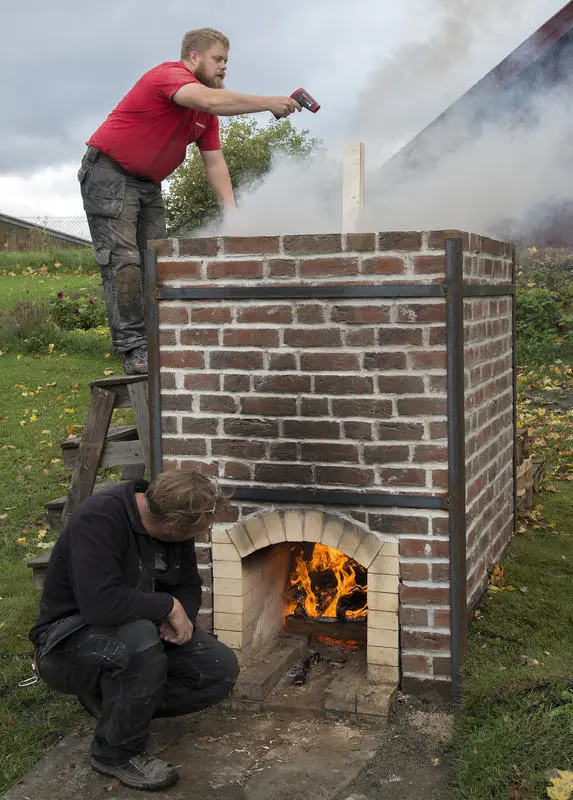 Mann i arbeidsbukser og rød T-skjorte står på trestige opp over kalkovnen og måler varmen, mens en svartkledd mann sitter på huk foran og ser inn i flammene.