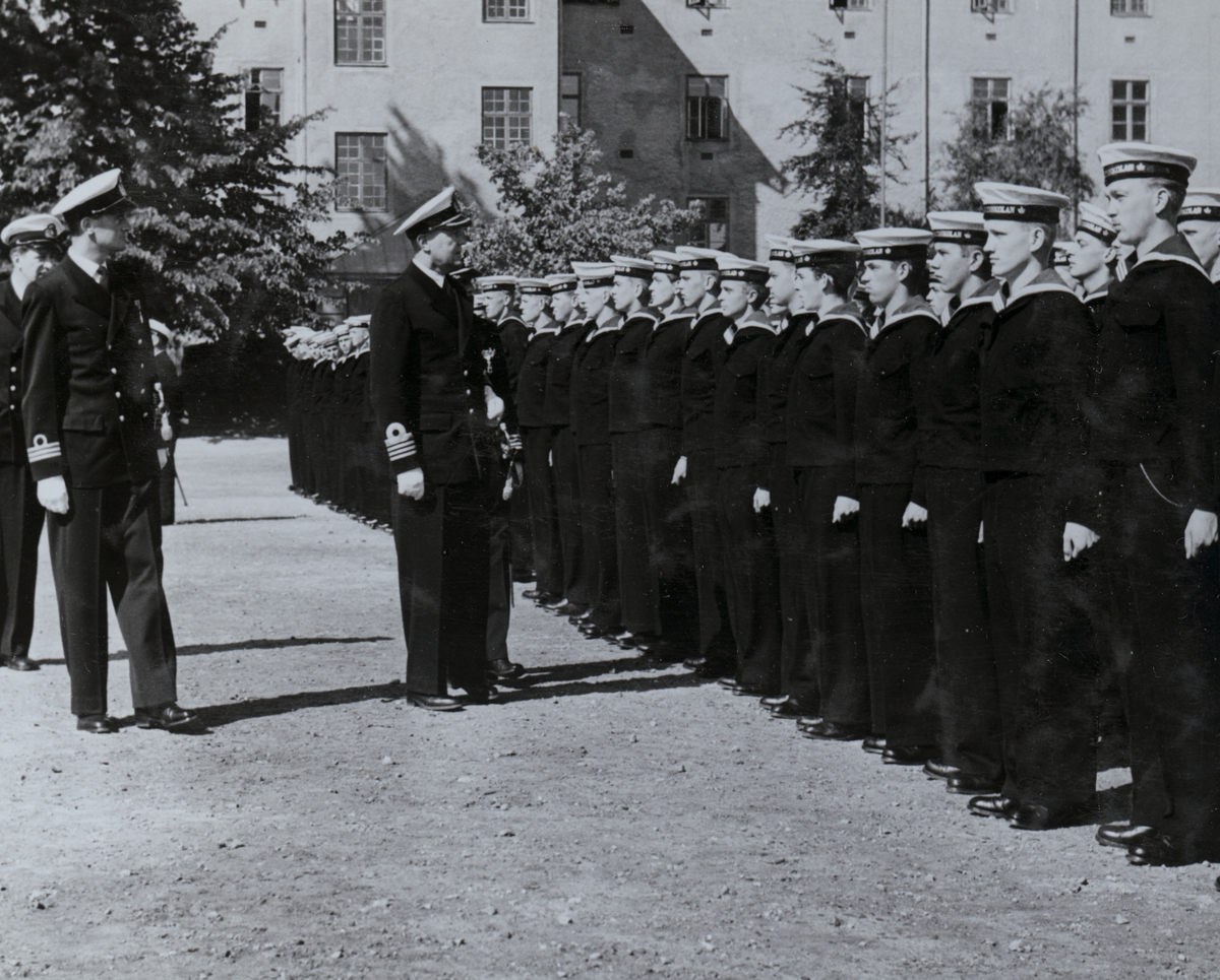 Uniformsklädda flottister står uppställda på rad inne på Sparres kaserngård. Framför dem går tre äldre uniformsklädda män med högre gradbeteckning, den främsta är kommendör Bong.