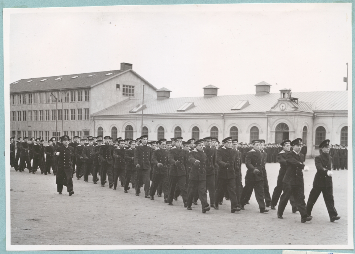 Uniformsklädda flottister marscherar över bataljon Sparres kaserngård. Längs med exercishusets putsade fasad med rundbågiga fönsteröppningar står grupper av uppställda flottister.