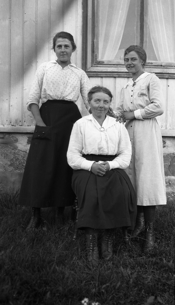 Tre kvinner i helfigur foran en husvegg. Foran sitter Anna Tømta (f1904), bak til høyre hennes søster Sigrid Tømta (f1900). Hun bak til venstre er ukjent. Bildet som er fra ca. 1920, er sannsynligvis fra Skullerud i Rossgutua på Skreia, som var Tømta-familiens hjemsted.
