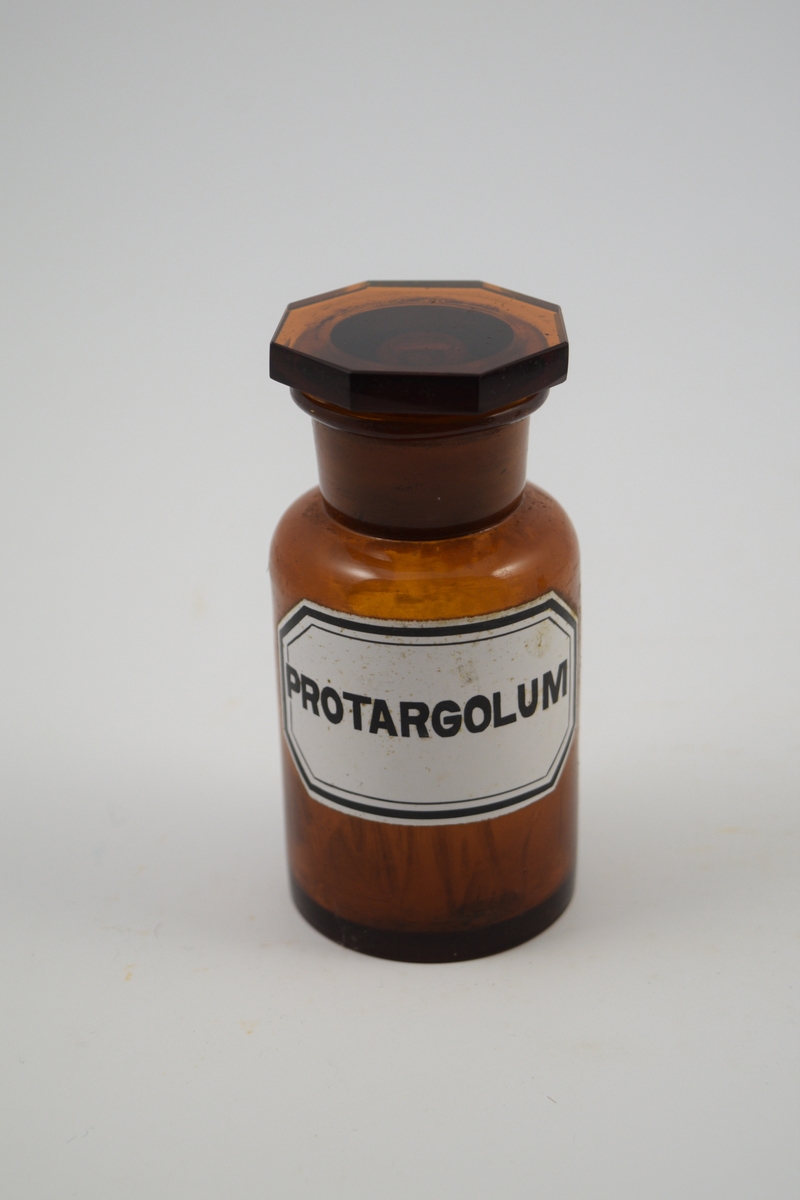Brun glasskrukke med brun propp. Hvit etikett med sort skrift. Brukt til oppbevaring av Protargolum (sølvproteinat), som tidligere ble brukt som nesedråper.
