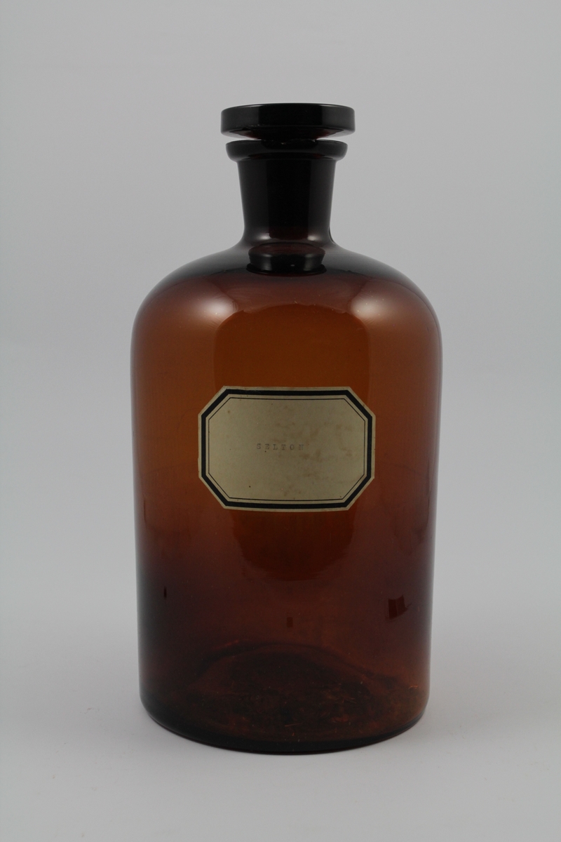 Brun glassflaske med smal hals, rommer 5 liter. Hvit merkelapp, sort skrift. Brukt til oppbevaring av løsninger.
