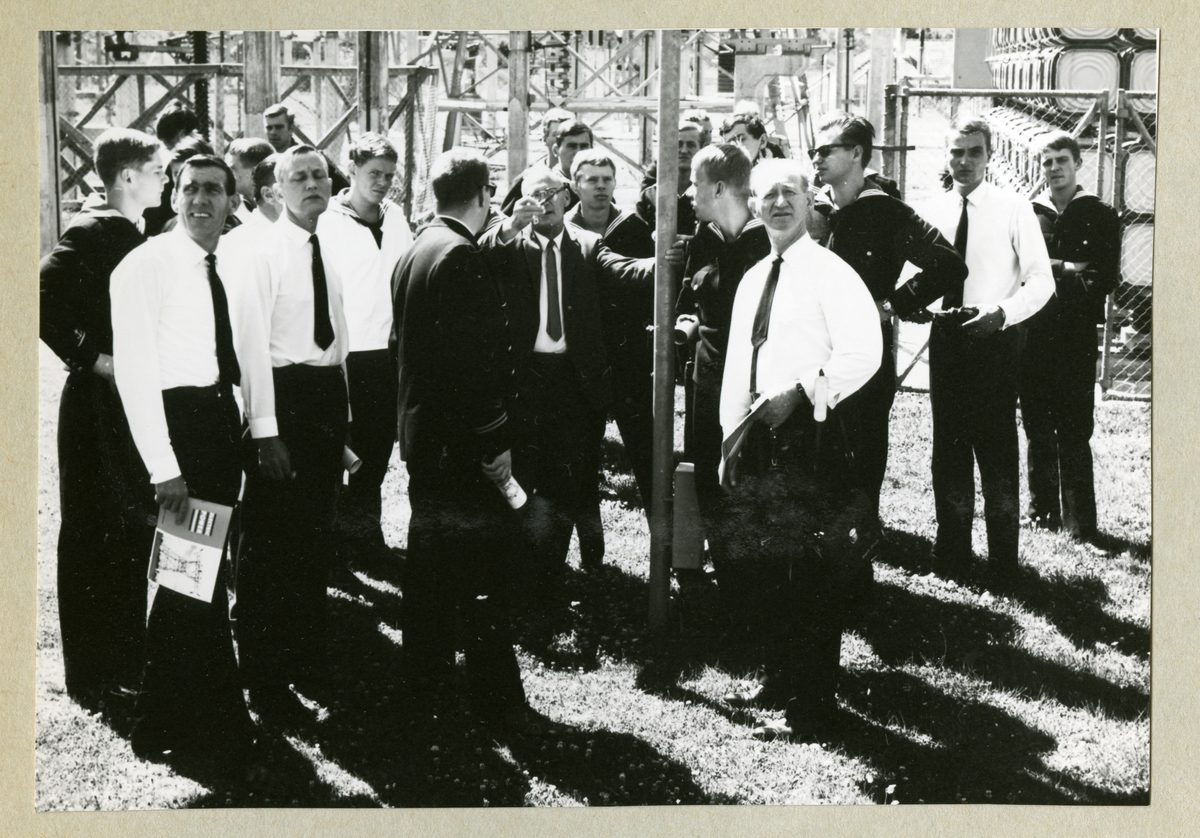 Bilden föreställer en grupp män som står samlade kring en man med rest hand. Alla besökande besättningsmedlemmar från Älvsnabben är klädda i uniform. En del har dock tagit av sig uniformskavajen eller bussarongen som tillfällig lättnad i klädseln.