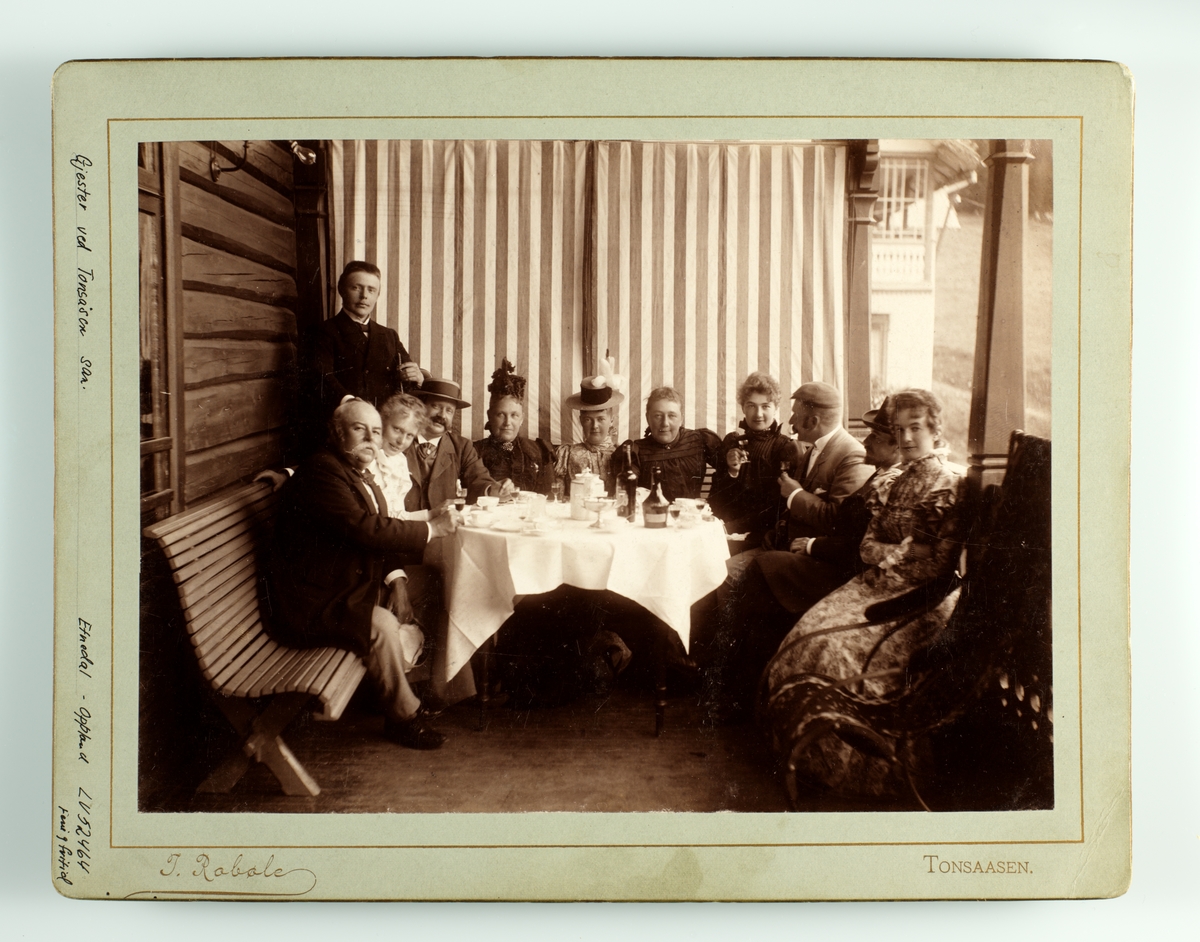Tonsåsen sanatorium, gruppe gjester samlet rundt dekket bord på åpen veranda. Kaffe- og alkoholservering på hvit duk. Trebenk mot veggen.
