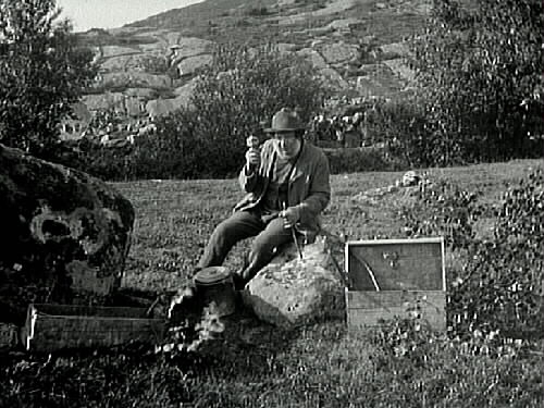 En stensprängare med skepparkrans sitter på en sten och drillar hål i den, omgiven av sina verktyg och redskap. I bakgrunden en bergssluttning. (Samme man på neg.nr. E5266.)
