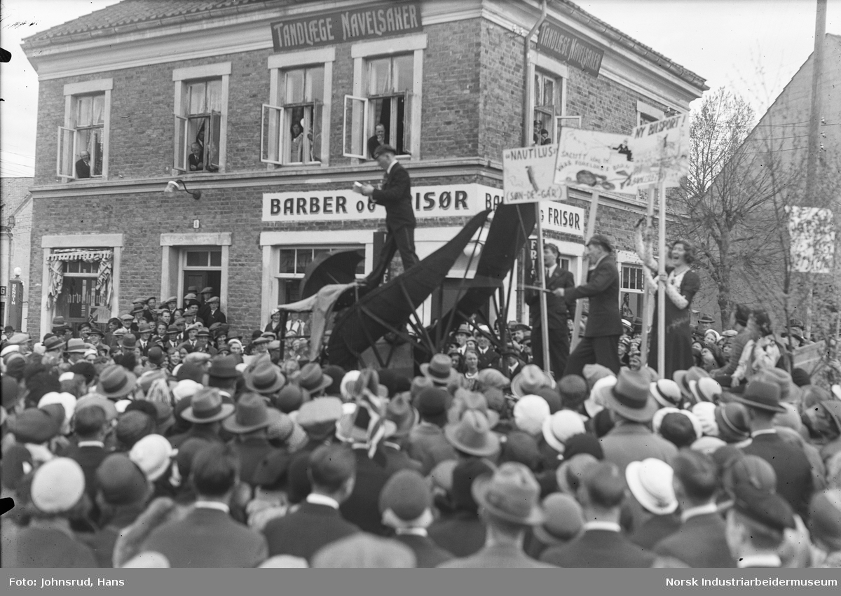 17. mai feiring 1933. Russ med lue og plakater holder tale til folkemengde på Notodden torg fra lasteplan. Folk i vinduene i hus i bakgrunnen. Butikklokale for barberer, frisør og tannlege Navelsaker.