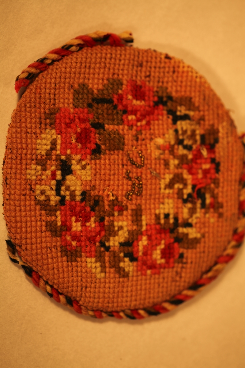 Rund pyntepute, brodert med blomstermønster på framsida, tvinnet band rundt puta, røde, gule og grønne farger. Brodert på baksida og på framsida.