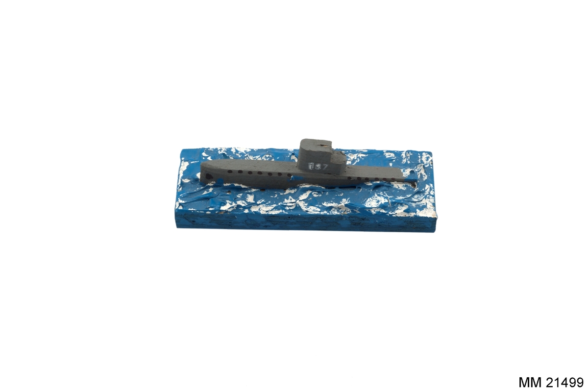 Ubåt på grund, tillverkad av trä, bestående av rektangulär platta, målad i blått med modellerade vågor. Ubåten i grått märkt: "137" på tornet. Papper på sidan av plattan: "KARLSKRONA".
Under platta text: "Aage Mazetti, Sturkö. Familjetillverkning".