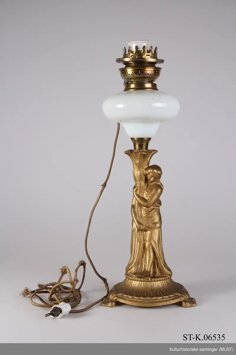 Skulpturell utformet lampefot med kvinnefigur som bærer en fakkel med en kuleformet beholder av hvitt glass.
Opprinnelig parafinlampe. Ombygd til elektrisk i senere tid.