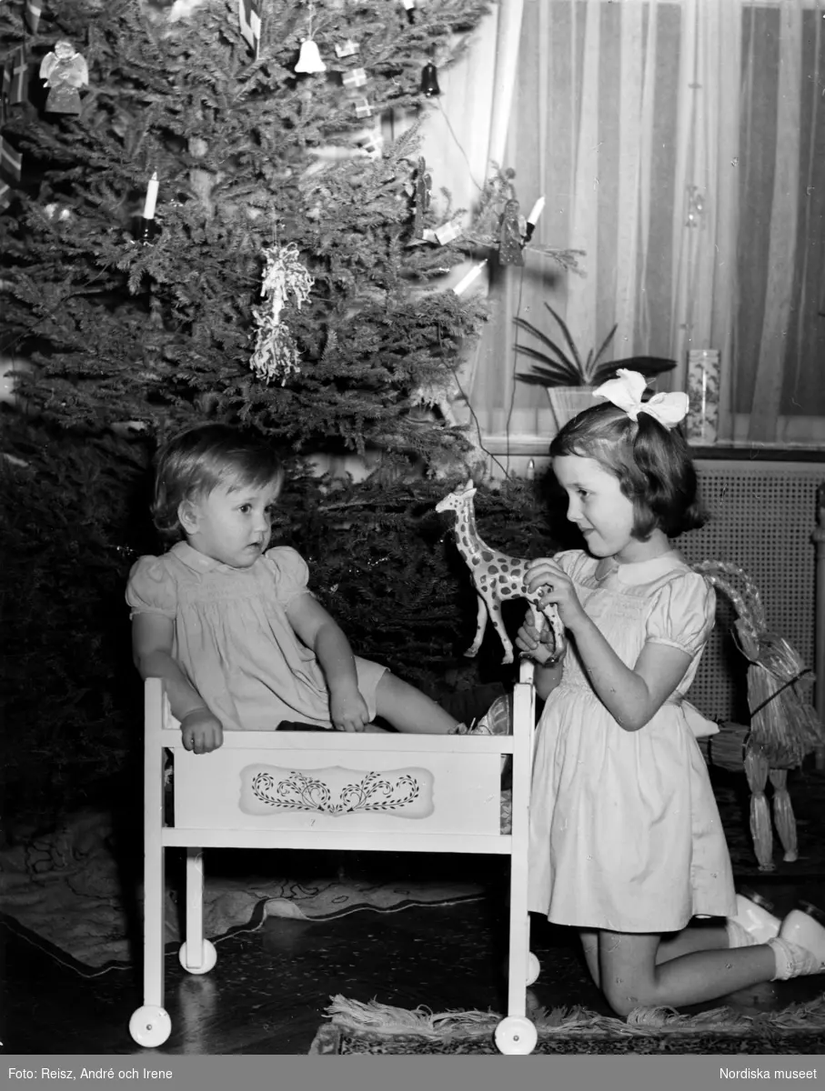 Två flickor vid julgran. Den yngre sitter i en docksäng på hjul, den äldre håller upp en leksaksgiraff