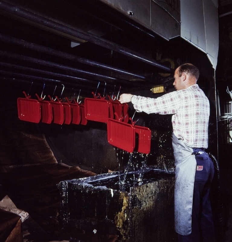 Arbeider henger "bagasjebrett" for trehjulssykler opp på et stativ før fornikling i forniklingsavdelingen til sykkelfabrikken til Jonas Øglænd AS på Sandnes, i fabrikkbygningen "Nr. 11".