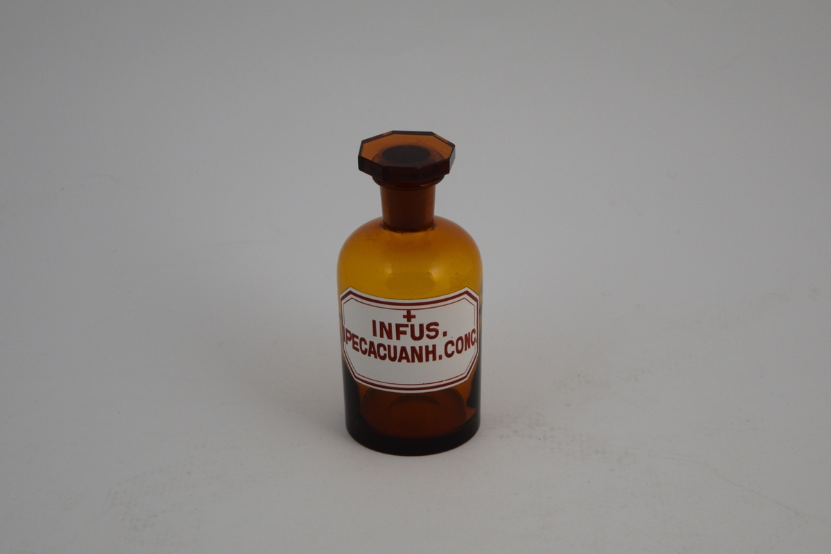Brun glassflaske, smal hals, åttekantet glasspropp, slipt. Påsatt hvit etikett med rød skrift og et rødt kors, noe som indikerer at innholdet er giftig. Krukken har inneholdt Ipecacuanha infus. (Brekkrot-infus).