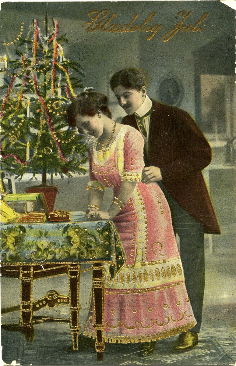 Julekort. Jule- og nyttårshilsen. Kurtisekort. Et festkledd par ved et bord med gaver og et pyntet juletre. Gulldekor på kortet. Stemplet 18.12.1916.