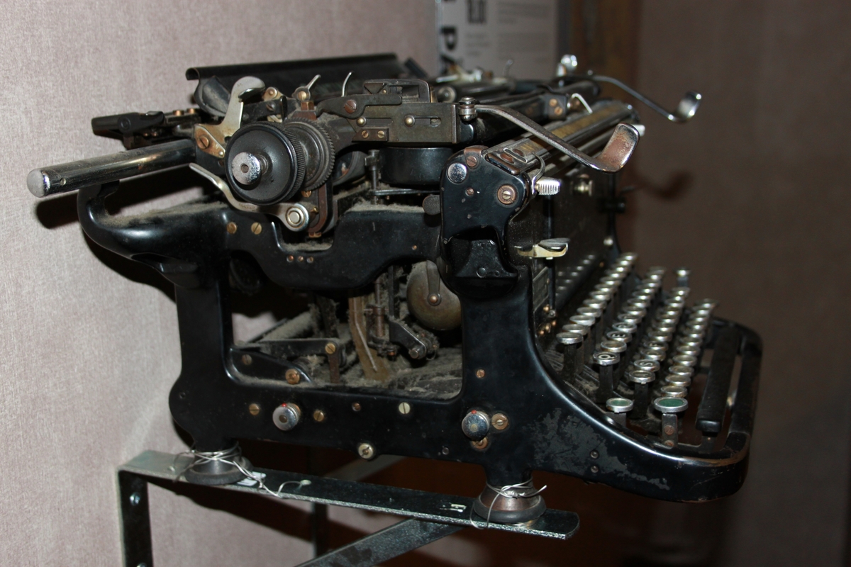 Rektangulær, mekanisk sort skrivemaskin med 49 tangenter på fire bein. Tangentene har sort bakgrunnsfarge.