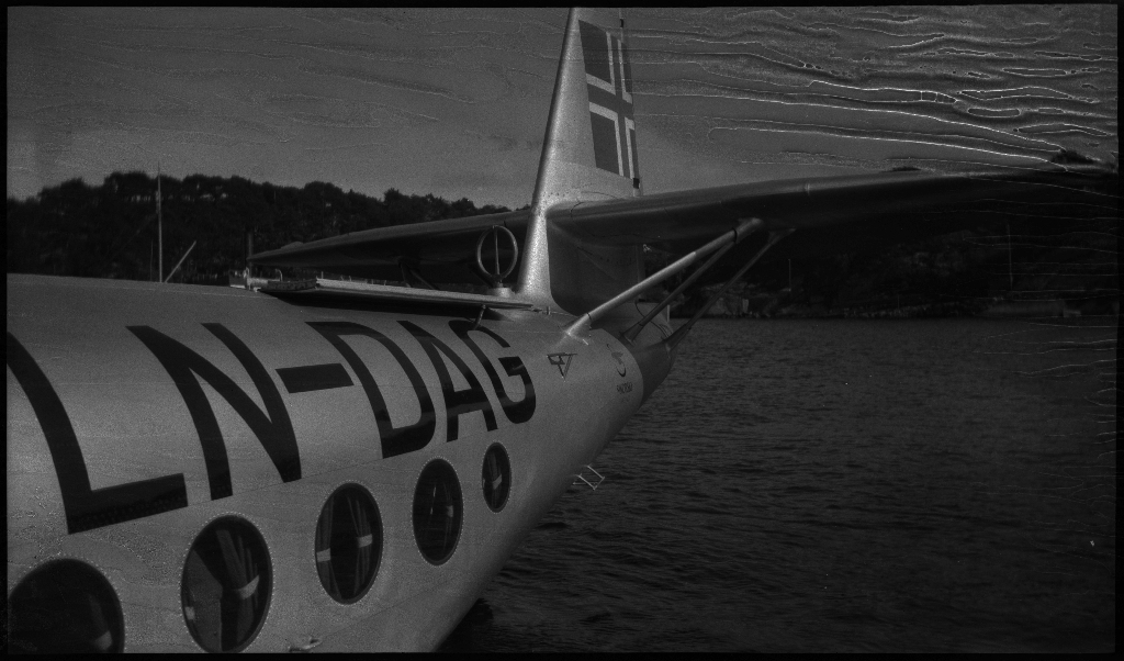 Bilder av Det Norske Luftfartsselskap sitt sjøfly (amfibie), "Valkyrien", som ligger på vannet. Registreringsnummeret, "LN-DAG", står på den bakre delen av flyet. En annen mann er med å besikte flyet.