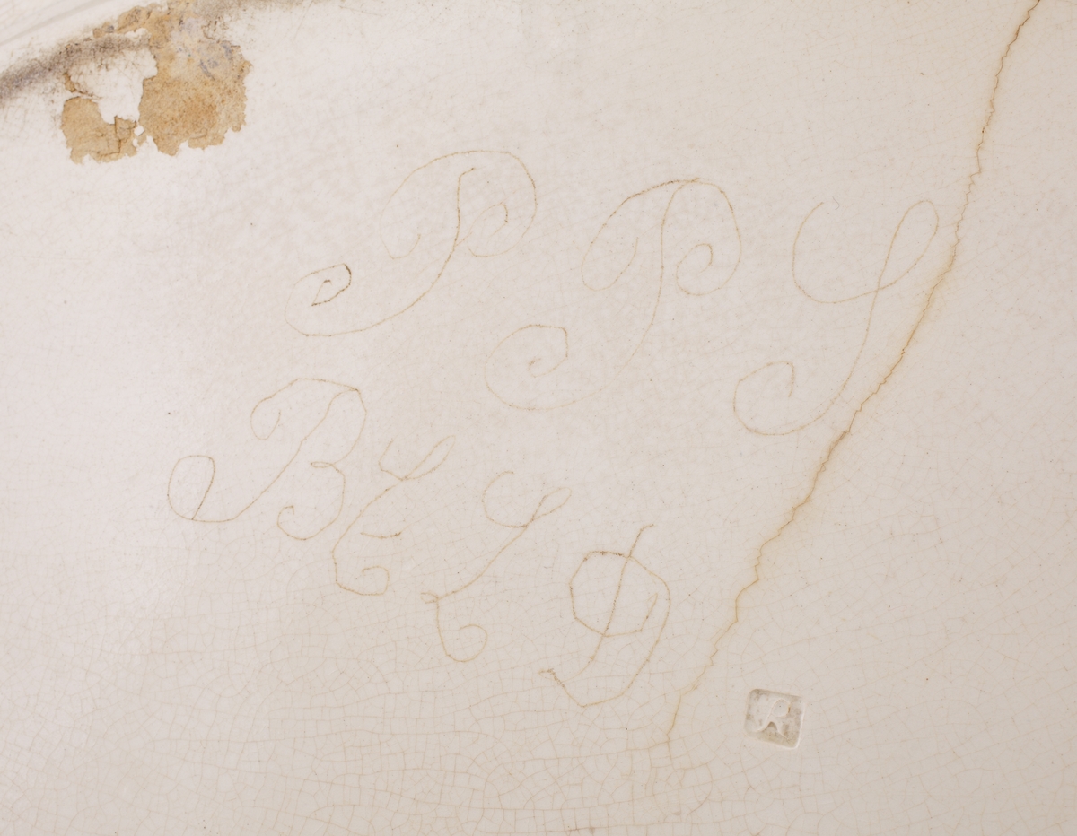 Uppläggningsfat av flintporslin.
Oval modell i beigevit glasyr, med vågig bräm i relief, s.k. feather edge. Ägarinitialer: "P.P.S., B.C.L.D." på undersidan av fatet. 
Stämpel på undersidan av fatet.