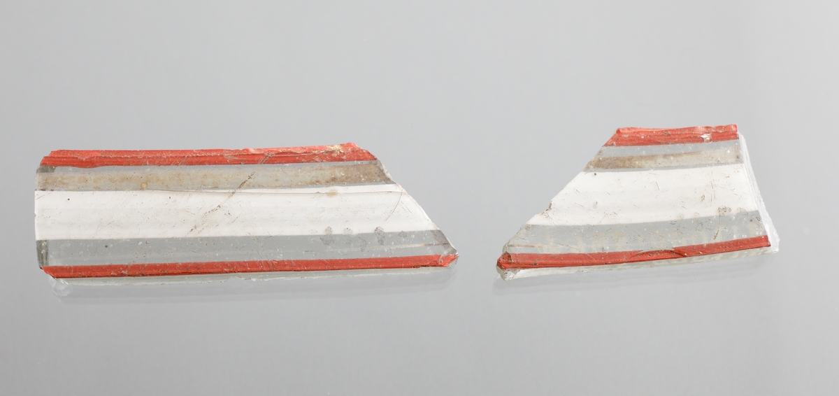 Två fragment av venetiansk glaspokal eller skål (a och b). Ofärgad glasmassa med inlagd dekor i form av två röda och en bredare vit tråd emellan dem.