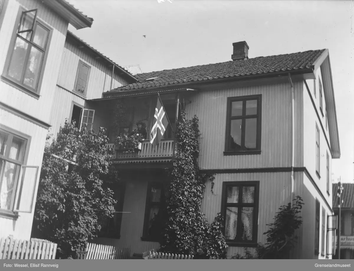 Stort bygårdshus med slyngplanter, kjellernedgang, ukjent sted. En kvinne står på balkongen i andre etasje, et flagg er hengt ut.