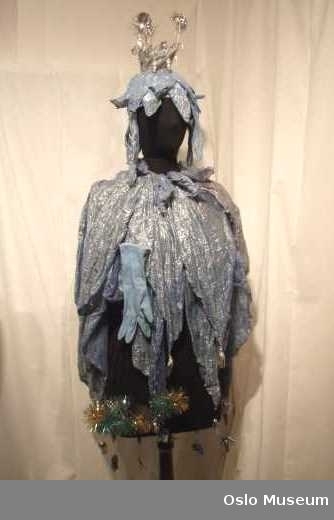 Cape, hodeplagg og 3 stk. enkelthansker. i lys blått/sølvstoff. Dekorert med dusker i juleglittermateriale samt perler på hodeplagg.