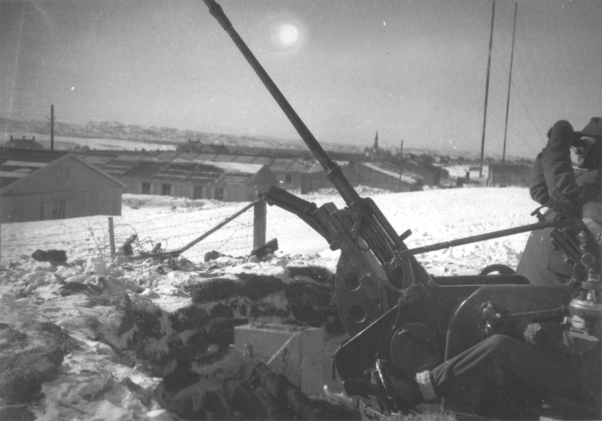 Vadsø. Vinter. Kanonstilling (20mm luftvern) med to soldater som sitter ved stillingen omtrent der Bergstien i Vadsø er idag. Vadsø kirke og tyske brakker sees i bakgrunnen.