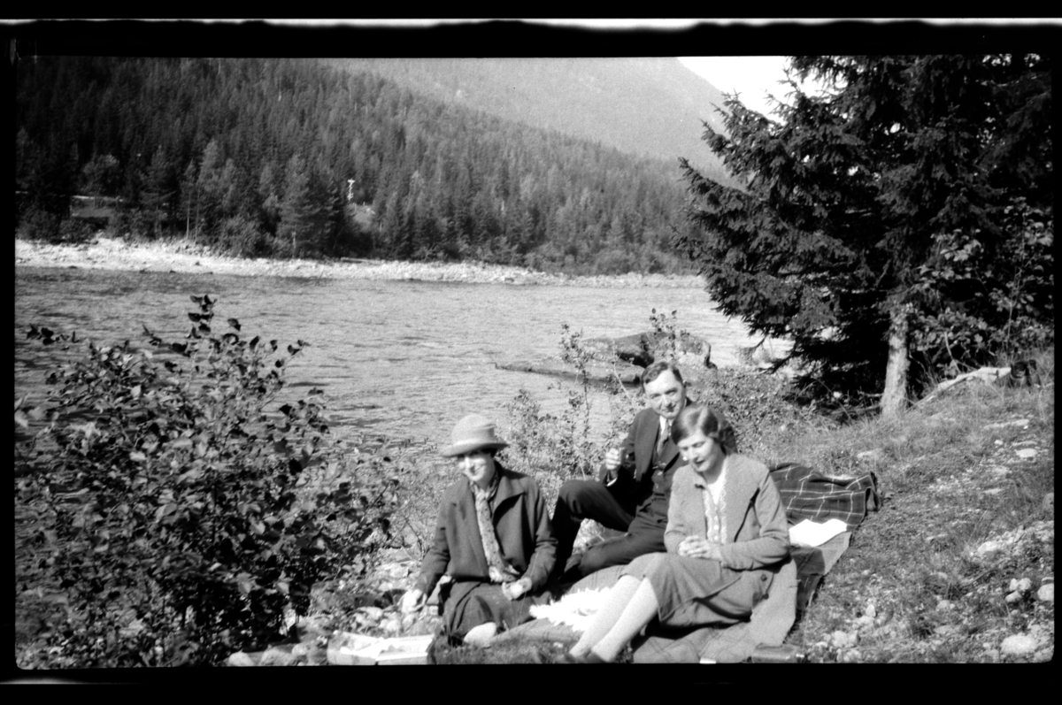 Ekteparet Agnes og Johan Buratti sitter sammen med  Hilda Sundt på et pledd ved en elv. Fotografert 1927.