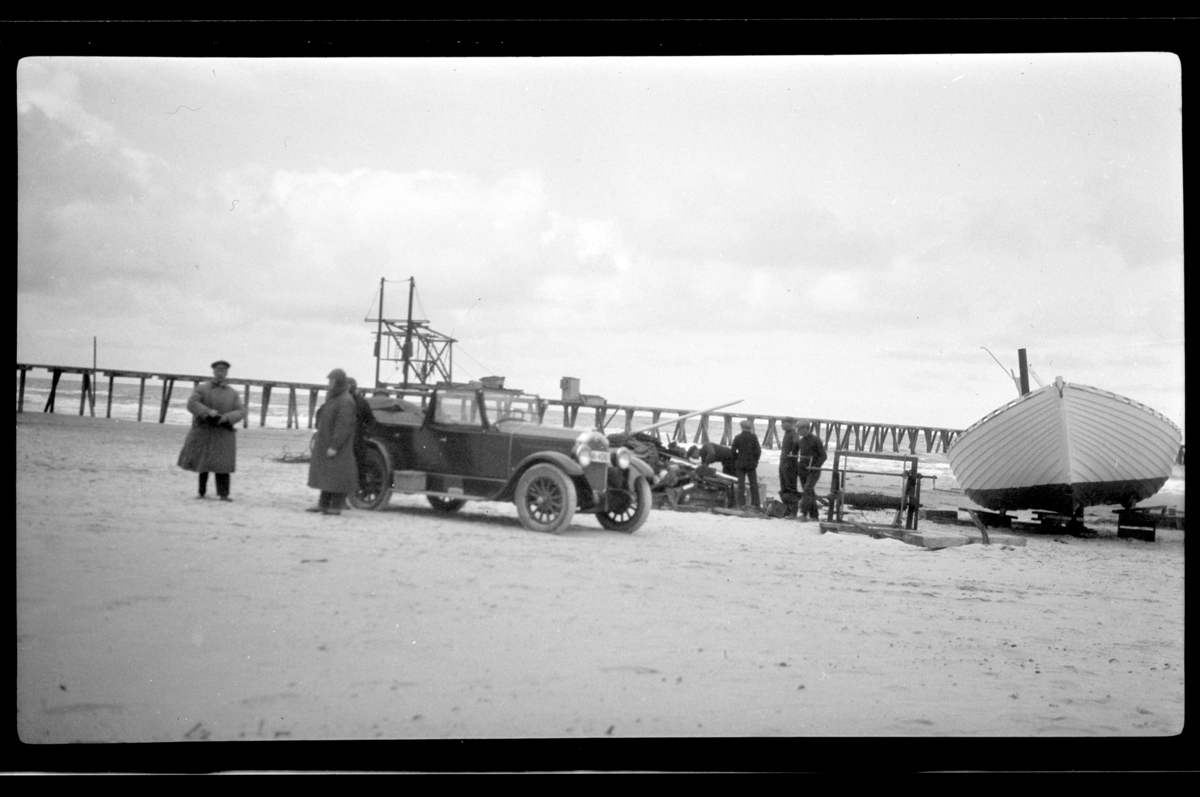 Til venstre står Rolf Sundt sr. ved siden av Buicken sammen med flere ukjente mennesker, i bakgrunn ligger en båt på standen i Skagen. Fotografert 1927.