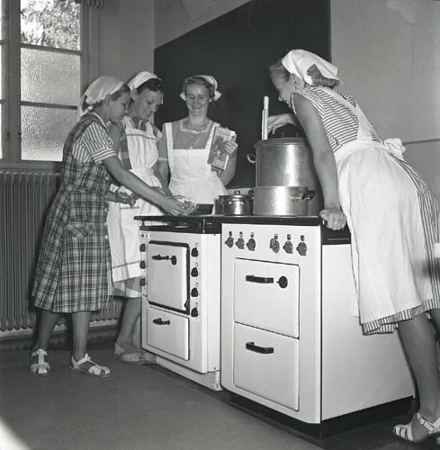 En kvinna och några flickor iklädda vita förkläden och sjaletter, står runt två spisar med grytor och kastruller. De ser ut att arbeta med konservering. Unga Odlare, Årsunda augusti 1950.
