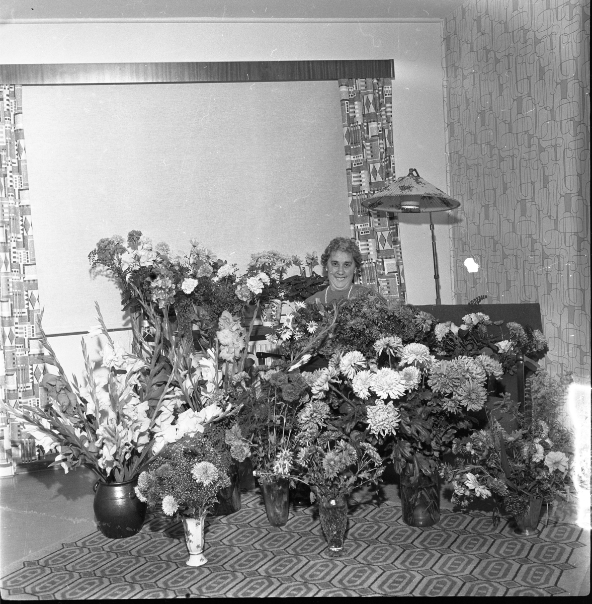 Signe Maria Johansson, Mellbyro, sitter i soffa framför ett stort fönster med nedrullad persienn. Framför henne på golvet står en stor mängd blomstervaser. Sannolikt födelsedag.