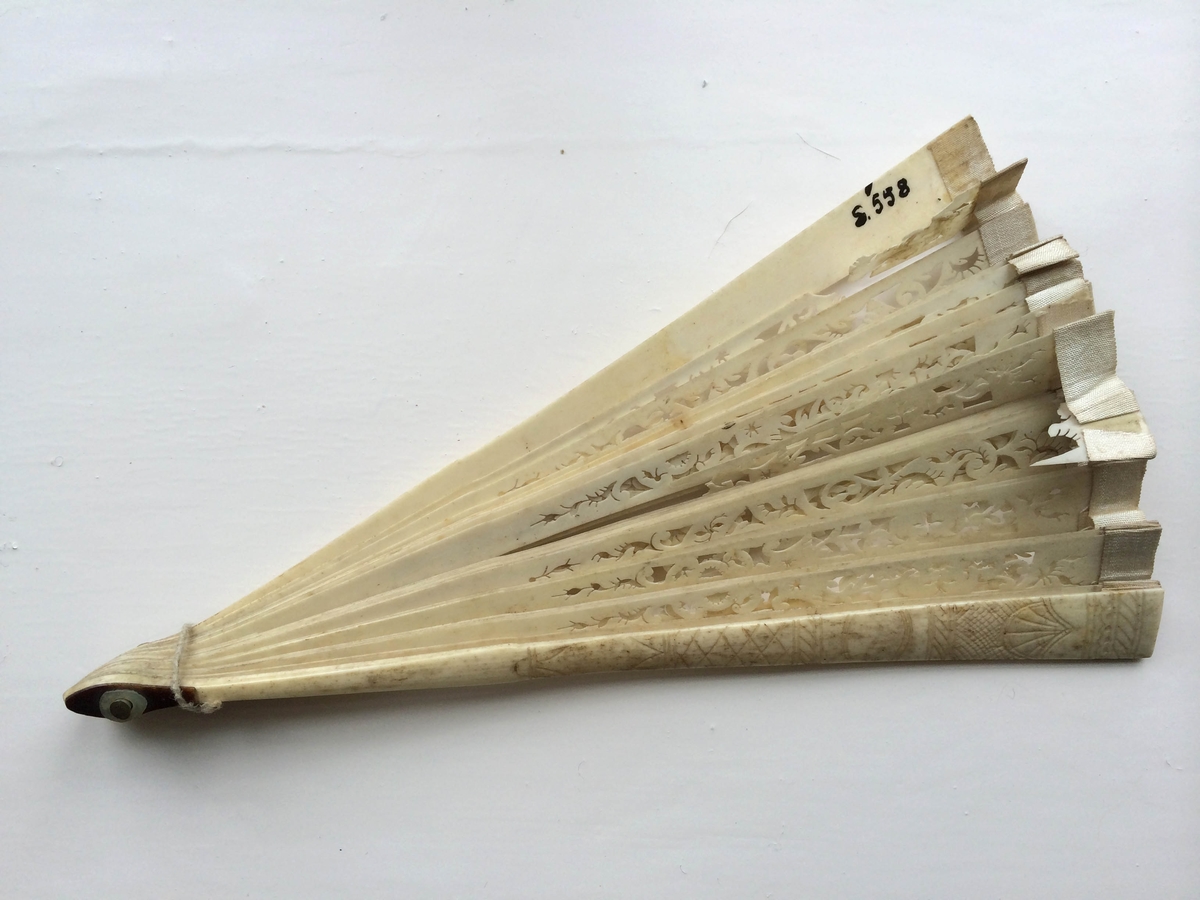 Vifte av ben, spiler og plate i ett, kantet med hvitt silkebånd øverst. Den del som danner platen er rikt utskåret, gjennombrutt i et rankemønster. Endespilene har et innrisset rute-, strek- og skjellmønster.