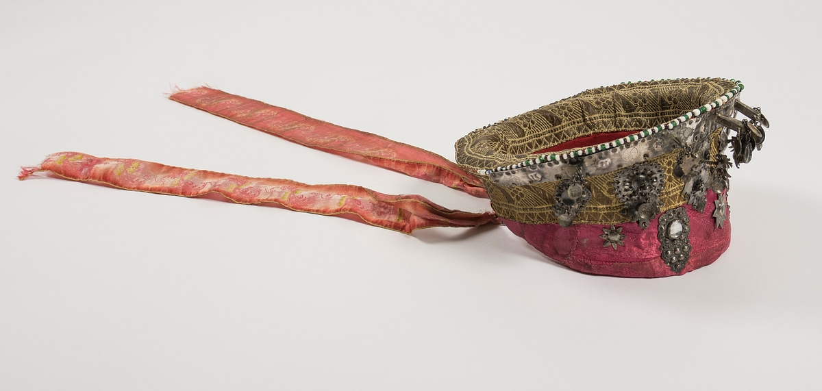 Valk, bruderull. Trøsken er i eit stivt materiale, del av den kan vera i never. Den er kledd med rosa silke.  Hjulet og øverste delen av trøsken er kledd med vevde metallband. Fòra på innsida med raudt toskaftvove ulltøy og ubleikt lin. Ytterst på hjulet oppe er det påsydd ei rad med kvite, svarte, grønne, blå og gule perler. Framme på trøsken er det festa ei smal sølvplate, denne har pålodda 6 stavar med ulike heng; bl.a. skåler, blank stein og lauv. På metallbandet under er det 6 sølvplater i ulike utformingar, (to og to er like). Under dette ( på den rosa tøyet) er det også to sølvplater.