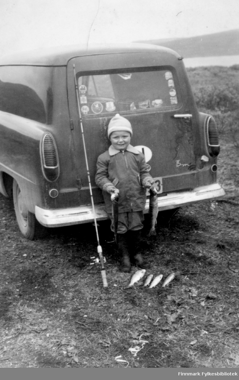 Barn med fiskefangst fotografert bak en Ford Taunus 15M varebil. Kfr 15M-logo står nederst til høyre bak på døren. Barnet står med fiskestang og flere fine ferskvannsfisker liggende på bakken. Barnet holder en fisk i hånden. Langs kanten av bilruta er det klistrert på mange merker med stedsnavn. Det var vanlig å kjøpe slike på bensinstasjoner når man var ute på ferieturer. Bildet kan være tatt i perioden 1955-1970. 

Familiealbum tilhørende familien Klemetsen. Utlånt av Trygve Klemetsen. Periode: 1930-1960.