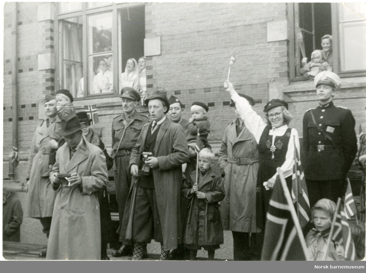 Festkledde mennesker med flagg utenfor Stavanger Sykehus 17. mai 1945. I vinduet til venstre er det flere sykepleiere. Jenta i vinduet til høyre er Astrid Lærdal (pikenavn) og hennes mor. Damen i bunad er hennes tante Hanna Marie Eskeland og mannen til høyre for henne i politiuniform er hennes onkel Odd Galtung Eskeland. To personer er i militær uniform. En person kikker på et kamera.