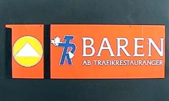 Rektangulär plastskylt med vit text på orange botten:
"TR BAREN
AB TRAFIKRESTAURANGER".
Symbolen "TR-pojken" i blått och vitt.