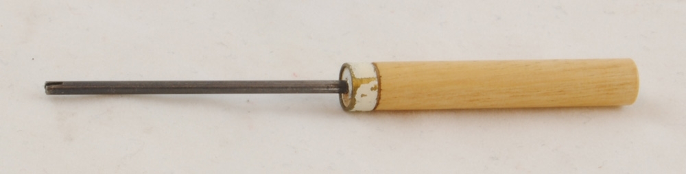 Verktyg med en metallpinne, 1,5x2 mm med en 2,5 mm djup skåra i änden. Verktyget har ett handtag av lackat trä med en metallring som är målad med vit färg, en markering att den tillhör teleavdelningen på SJ-skolan.