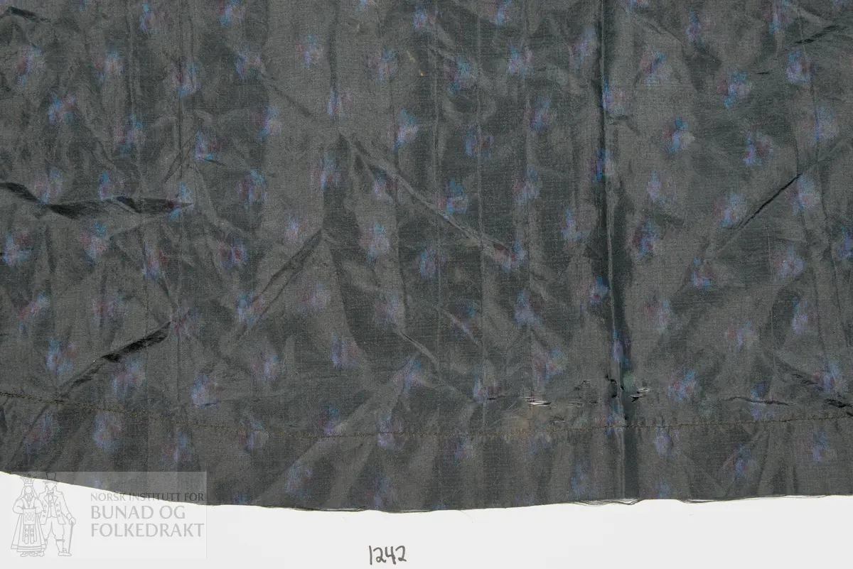 Forkle, laga i svart silketaft med trykt mønster i blått, mellomblått og lilla. Lett utskrådd, samansett av 3 høgder. Maskinsaum. Svart silkeband til knyting. Faldelagt med fire faldar oppe.