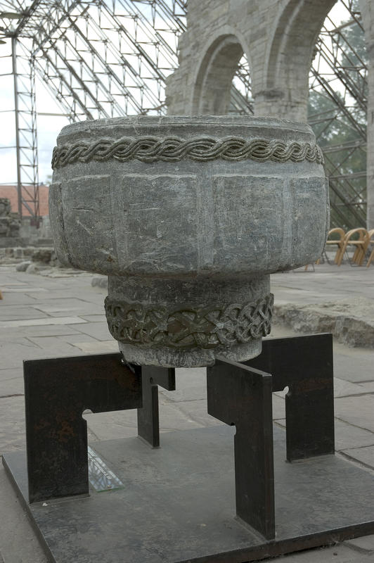 Døpefont fra middelalderen, i kleberstein med flettebord øverst, står på metallstand i Hamardomen, med domkirkeruinen i bakgrunnen.