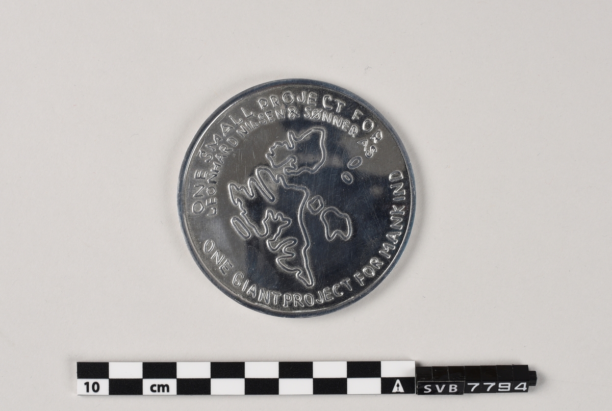 Skive av støpt metall med presset mønster og tekst på begge sider.
Medaljen ligger på en skumpute i en svart kunststoffeske med lokk .