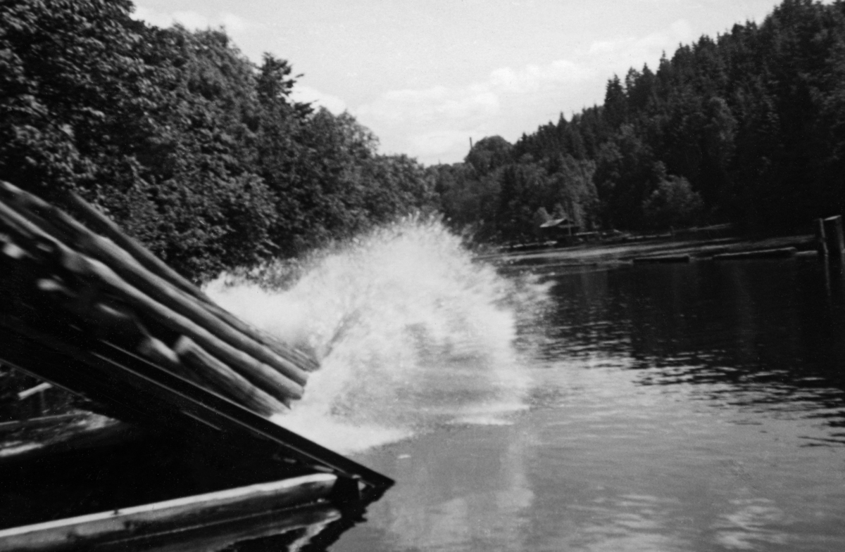 Utislag av buntet tømmer i det kanaliserte Haldenvassdraget.  Fotografiet er tatt i samband med Norsk forstmannsforenings utferd i denne regionen 10. -11. juni 1949.  Det viser en tømmerbunt som sklir nedover på et skråplan fra en utislagsplass for lastebil (jfr. SJF-F. 006109), og når vannspeilet med et plask.  Lengre ute i vannet, til høyre i bildet, skimter vi en påle, samt en del fløtingstømmer.  Langs denne delen av vannveien vokste det mye lauvskog.  Etter 2. verdenskrig ble bruk av lastebiler i tømmertransporten stadig vanligere.  Ved de store fløtingsvassdragene ble det opparbeidet velteplasser, der tømmer som var hentet langs de mange nye skogsbilvegene og kjørt til hovedvassdragene og slått uti, slik fotografiet viser.  Haldenvassdragets fellesfløtningsforening later til å ha vært tidlig ute med slike anlegg. 