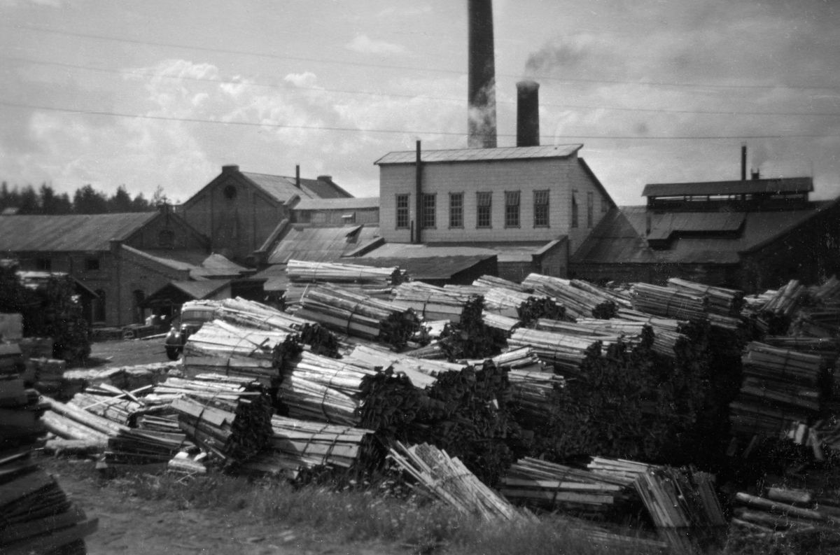 Bildet er tatt ute på fabrikktomta fra øst mot fabrikkbygningene på Klevfos Cellulose- & Papirfabrik.  Honlageret i forgrunnen vitner om at bedriften økte bruken av dette avfallsproduktet fra sagbrukene.  Fabrikken brukte cirka 5000 kubikkmeter bakhon tidlig i 1960-årene. 
Det er to fabrikkpier i drift. Den bakre pipa, som det kommer ut røyk fra, er tilknyttet fyrhuset mens den runde skrosteinen er for sodahuset. Sodahuspipa var ny i november 1956. Den ble tatt i bruk 18. november 1956. En kort periode høsten 1956 var fabrikken utstyrt med tre piper, en for fyrhus og to for sodahus. Den gamle firkantete sodahusskorsteinen ble revet 26. novmeber 1956.  Like foran sodahuspipa ser en det karakteristiske overbygget over det nye lutinndampingsanlegget. Lutinndampingsanlegget ble innkjøpt brukt fra Torp Brug i 1955. I august 1956 kom prøvedriften av anlegget i gang. Bygget over anlegget som kom på plass i 1955/1956 hadde først bordkledning. Denne bordkledningn ble seinere påført vedlikholdsfire eternittplater. Da dette bildet ble tatt var veggene kledt med vedlikeholdsfrie eternittplater. 

Bildet er udatert, men det grunn til å tro at det er tatt en gang i tidsrommet 1959-1963. Det er derfor grunn til å betrakte bildet som en illustrasjon på hvordan fabrikkanlegget så ut omkring 1960.  Løten. Ådalsbruk.