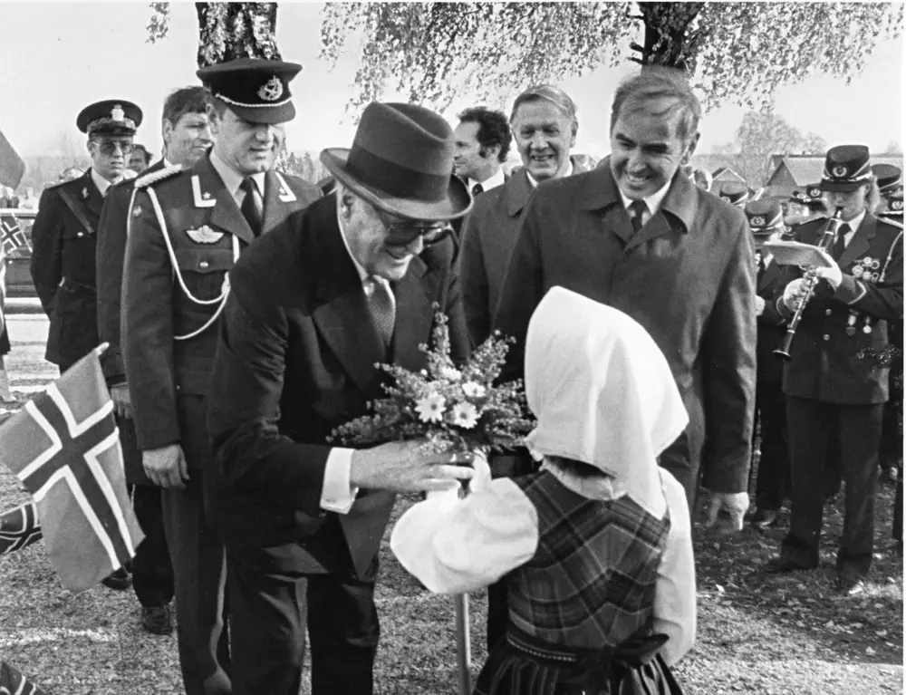 En mann med hatt mottar blomster av en ung jente. Omkring dem er mennesker i penklær og norskeflagg.